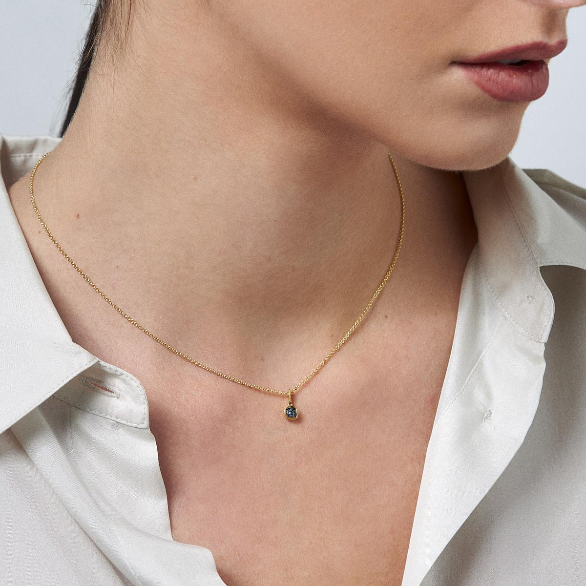 A close-up of a woman wearing a delicate gold chain necklace with a small dark blue teardrop pendant, paired with a white collared shirt against a light gray background.