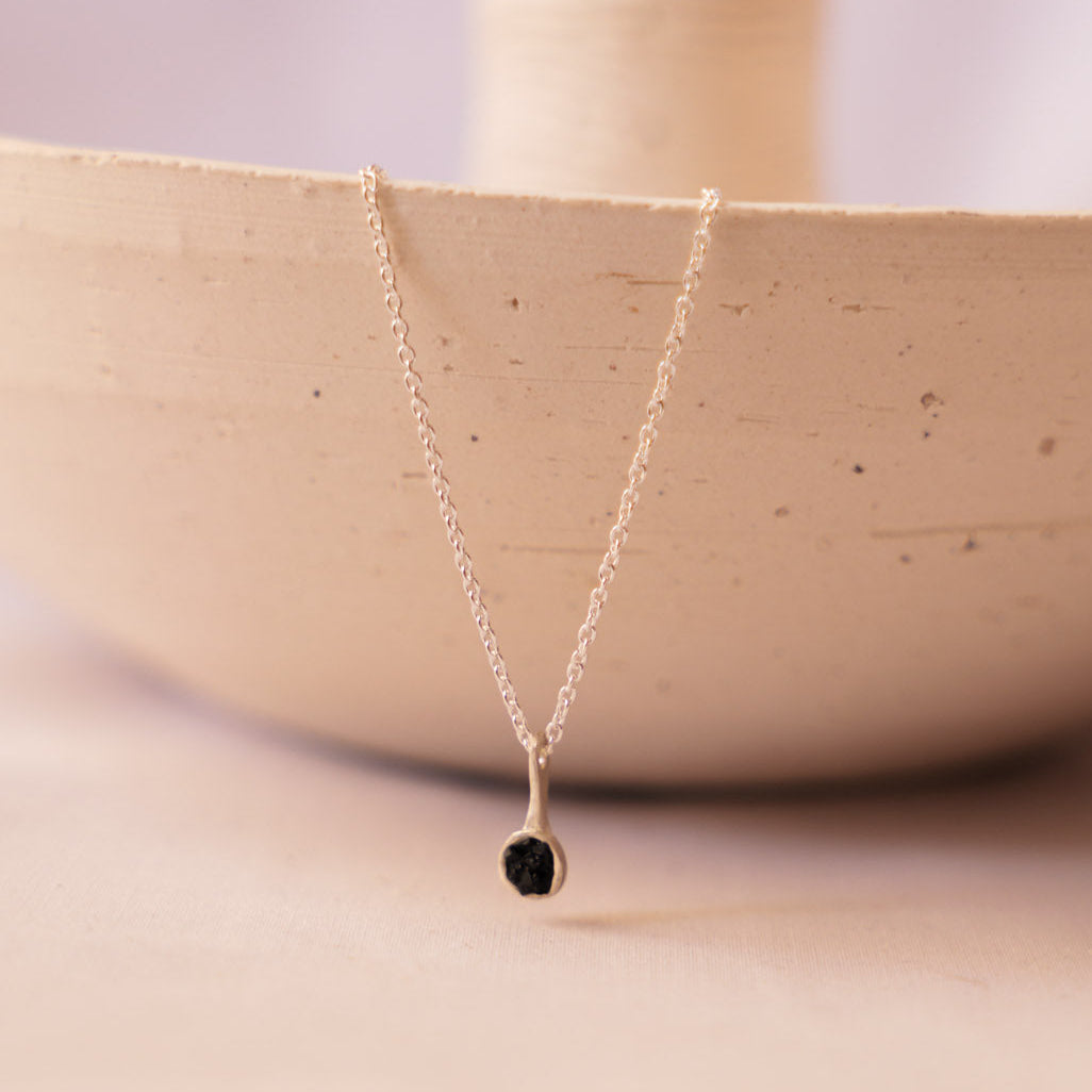 A delicate silver chain necklace with tiny fragments of hematite pendant, draped over the rim of a beige ceramic bowl on a soft beige background.
