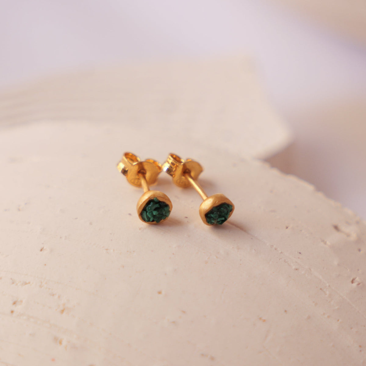 A pair of gold stud earrings with small green fragments of malachite resting on a textured beige ceramic plate against a soft white background.