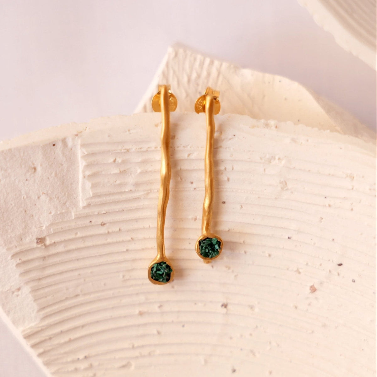 A pair of gold dangle earrings with tiny fragments of malachite hanging from a light beige ceramic dish on a soft neutral background.