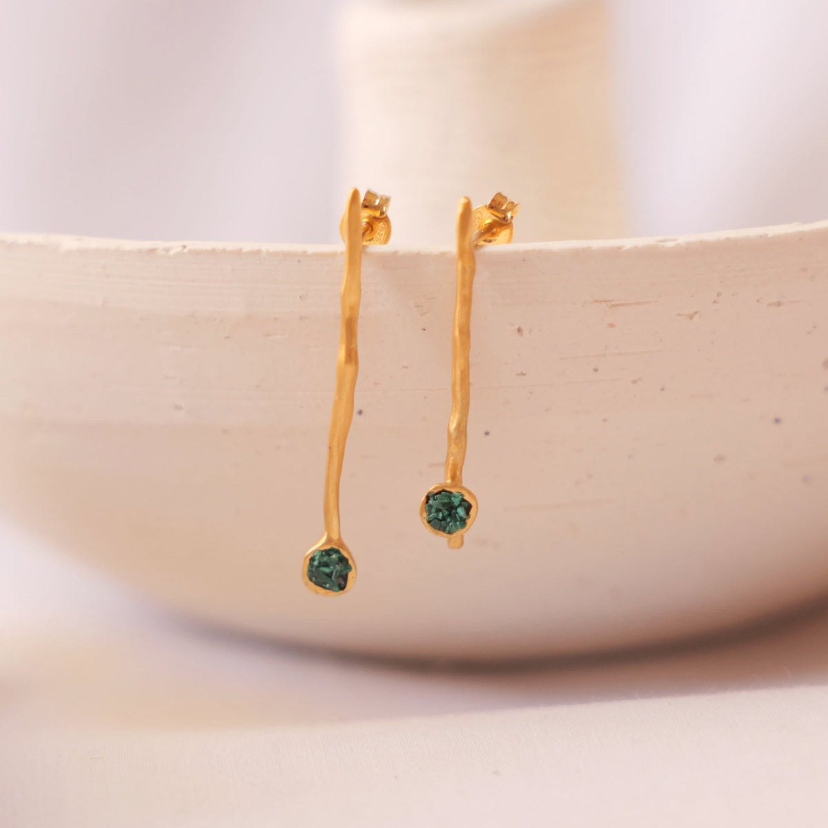 A pair of gold dangle earrings with tiny fragments of malachite hanging from a light beige ceramic dish on a soft neutral background.