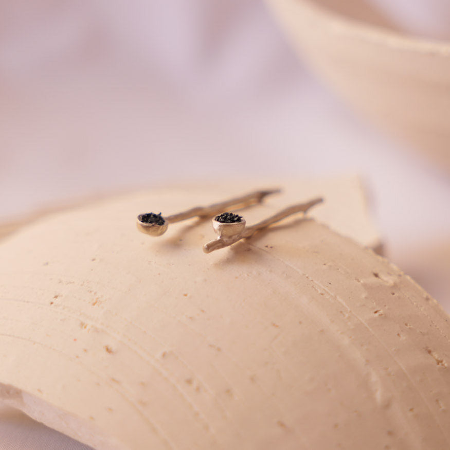 A pair of small gold post earrings with dark round stones, displayed on a textured beige ceramic dish against a soft, warm off-white background.