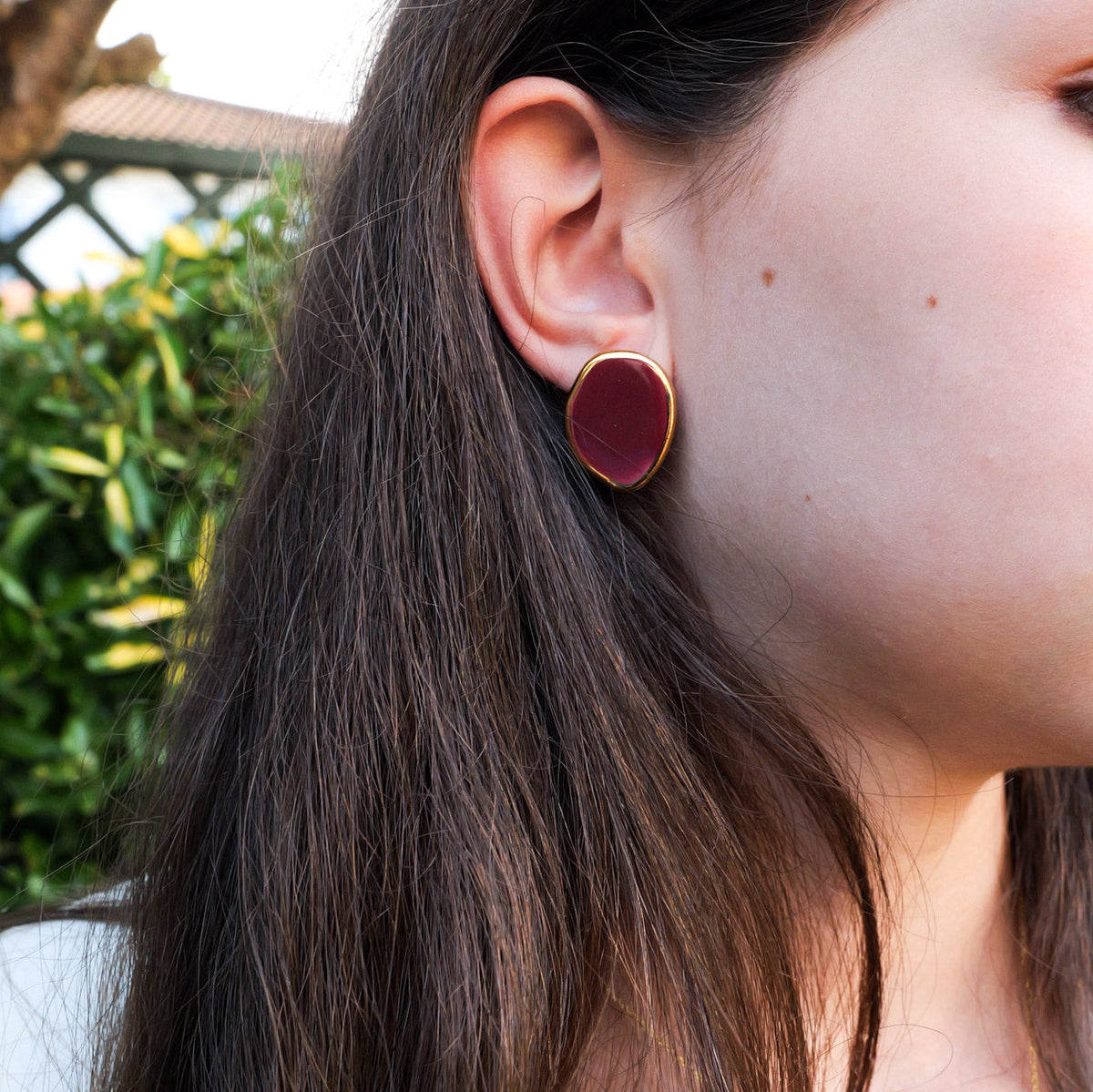 A close-up side profile of a woman wearing a gold-framed oval earring with a deep burgundy ceramic center, set against a blurred outdoor background of green foliage.