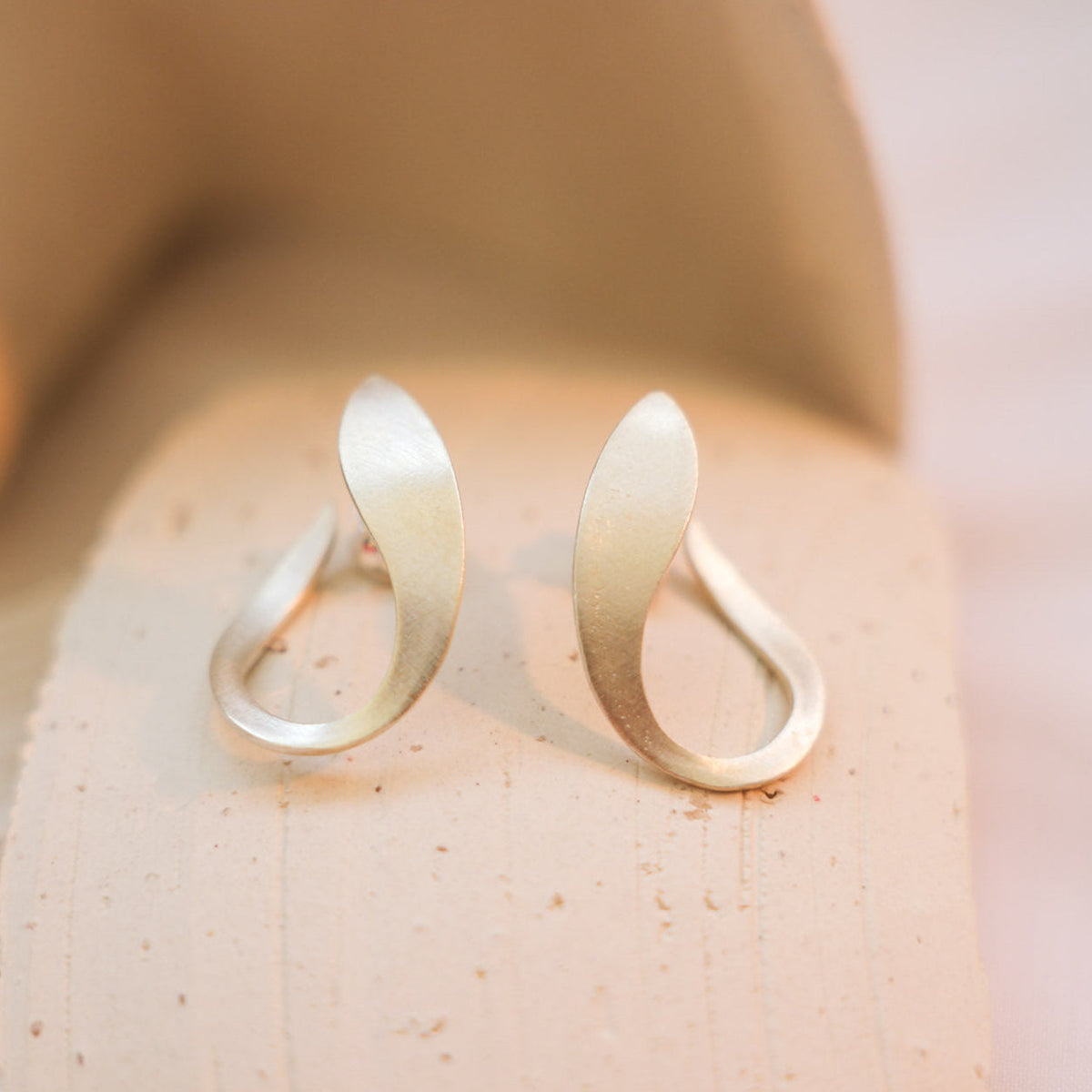A pair of sterling silver minimalist silver earrings with a curved, open teardrop design, resting on a textured beige stone surface.