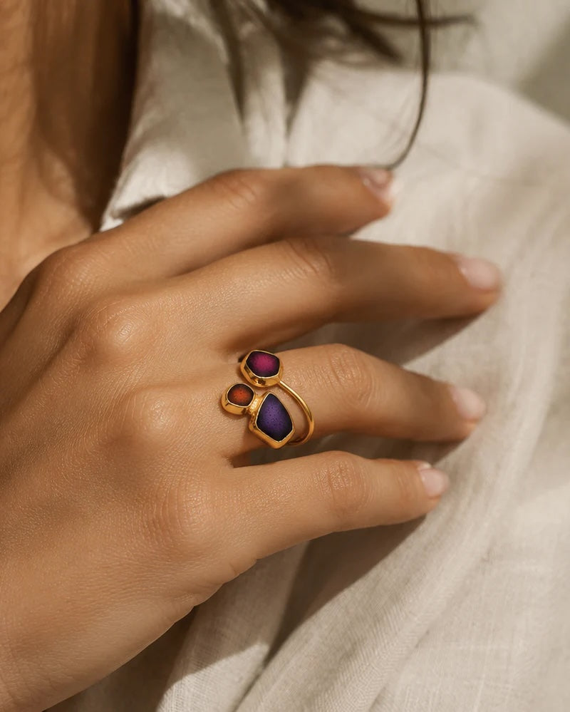 A gold ring with three marquise-cut purple and magenta stones, worn on a person's finger against a soft beige linen background.