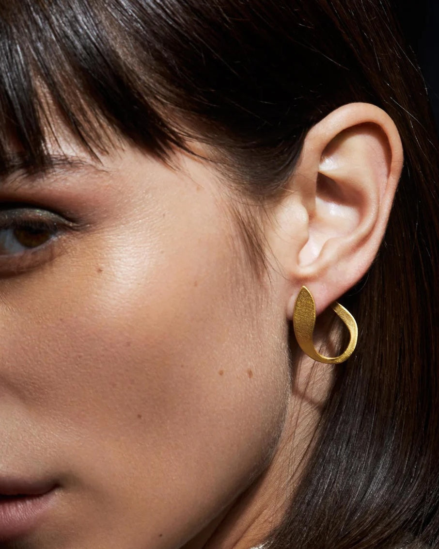 Close-up of sculptural matte gold-plated earrings with a softly curved organic silhouette worn on the ear.