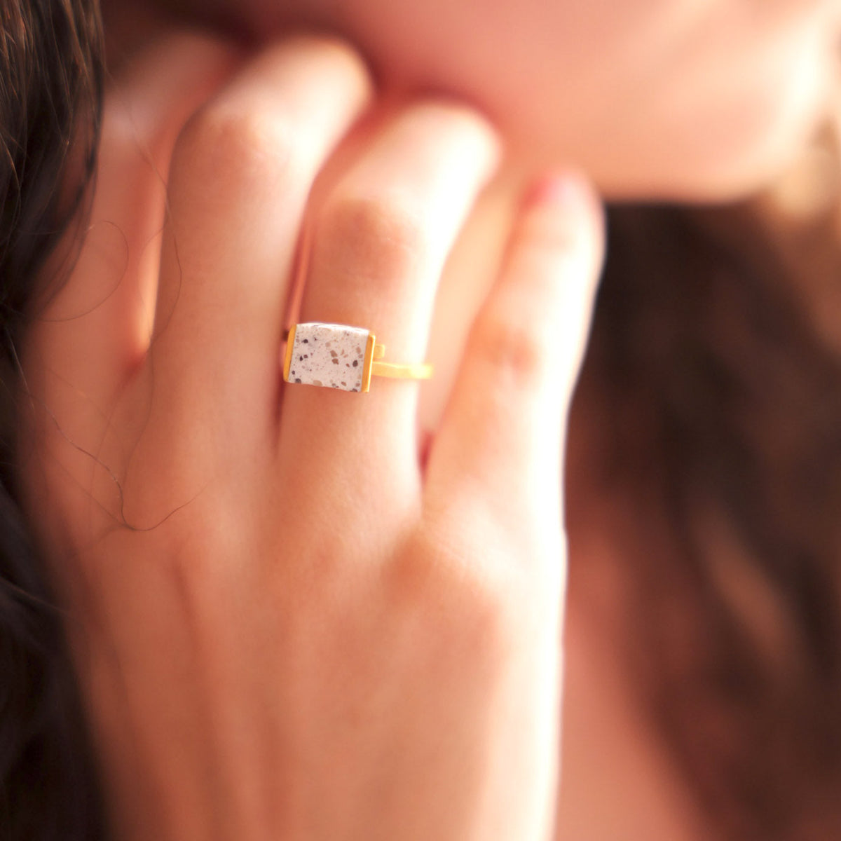 A close-up lifestyle shot of a person's hand wearing a gold ring with a rectangular whiten terrazzo stone containing dark speckles.