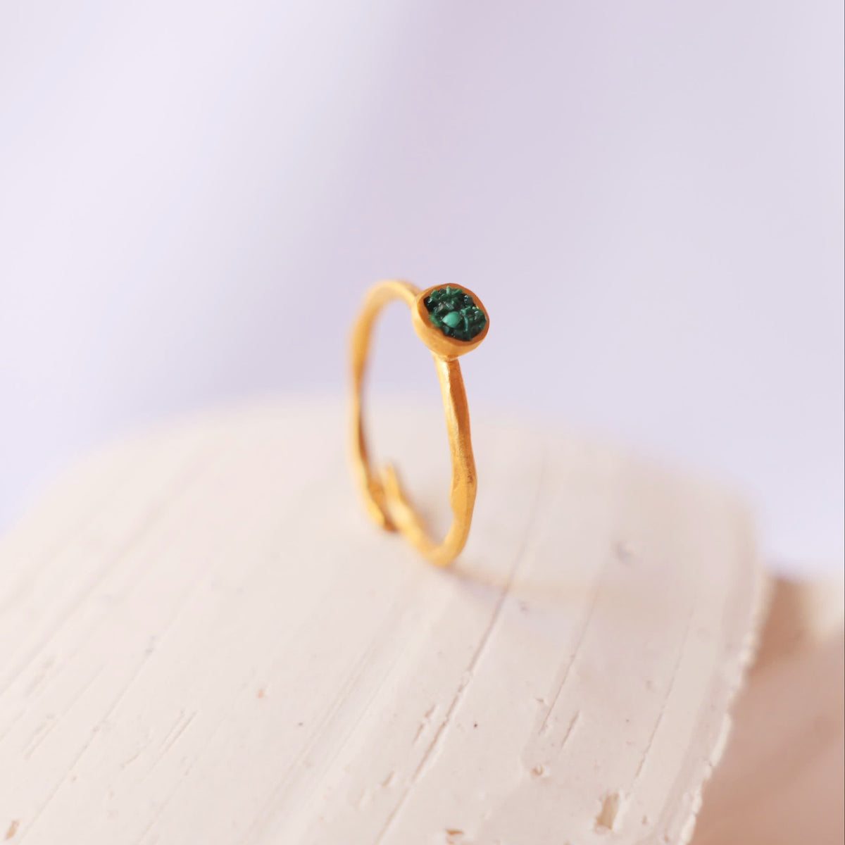 A delicate gold ring with a small, round green gemstone, displayed on a textured beige stone block against a soft, light background.