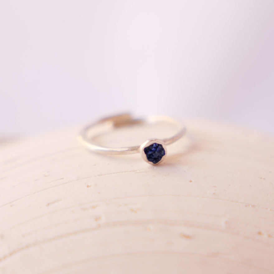 A delicate silver ring with a small, faceted dark blue gemstone, resting on a textured beige ceramic dish against a soft, light background.