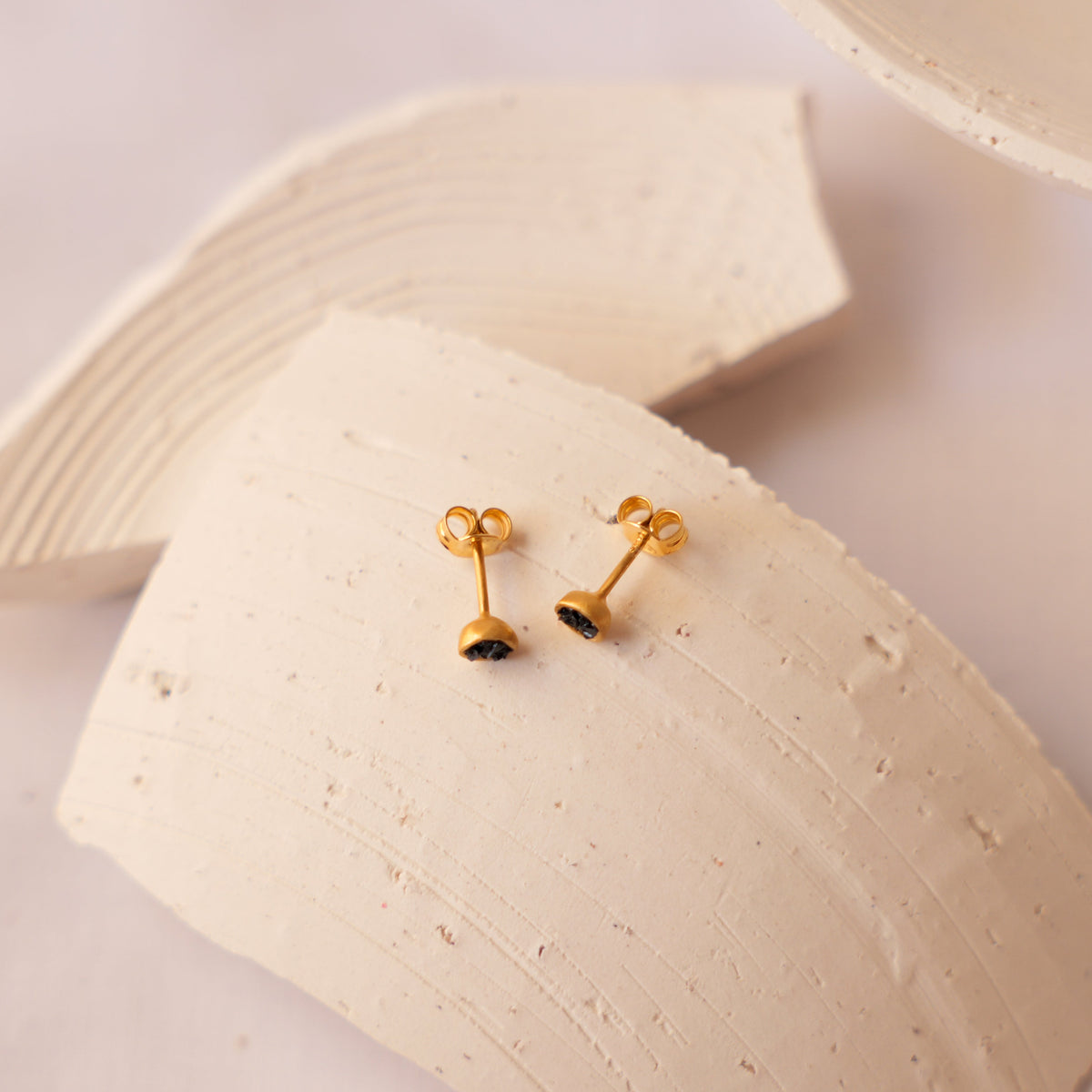A pair of small gold stud earrings with tiny fragments of semi precious stones, displayed on a textured beige ceramic prop against a light background.