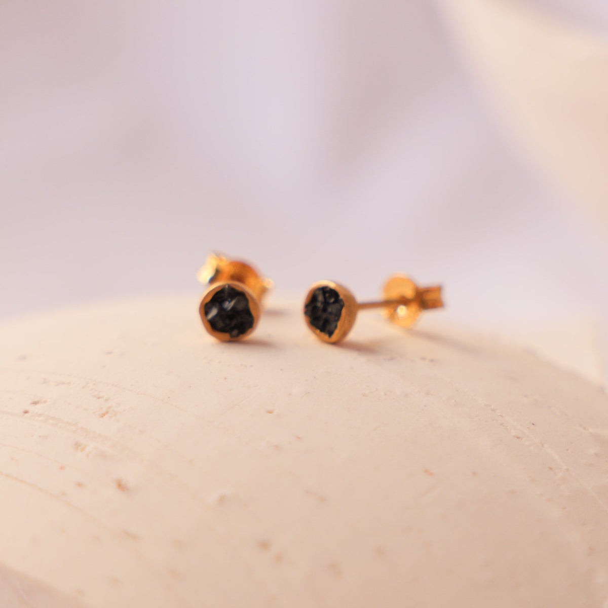 A pair of gold stud earrings with small green fragments of hematite resting on a textured beige ceramic plate against a soft white background.
