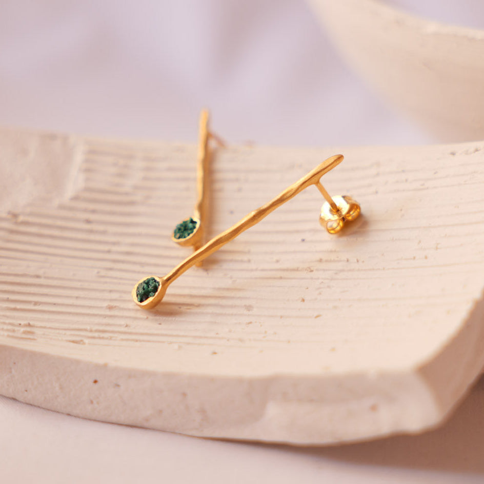 Gold branch-shaped stud earrings with small green fragments of malachite displayed on a textured ceramic dish against a soft neutral background.