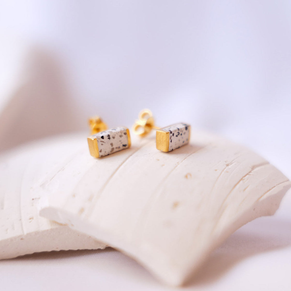 A pair of small gold stud earrings with rectangular white terrazzo stones with dark speckles, resting on a textured white ceramic tile against a soft, out-of-focus light background.