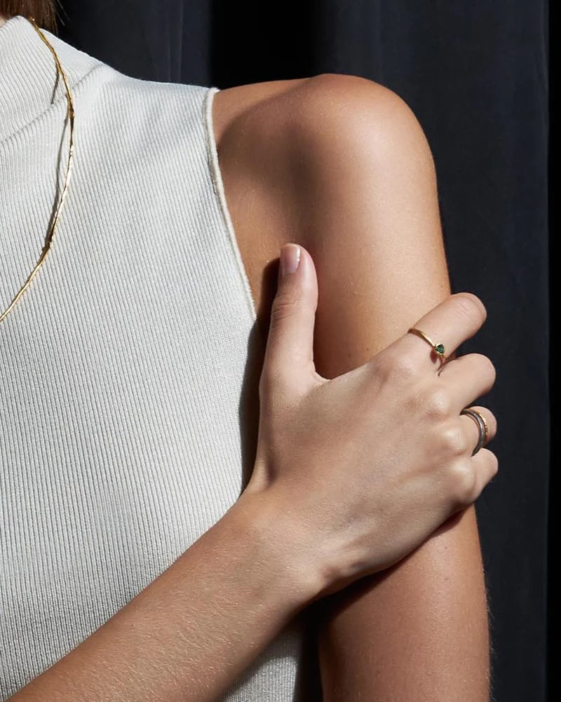 A close-up lifestyle shot of a model's upper arm and hand wearing delicate gold rings, including a thin band ring with a small green stone and a textured band ring.