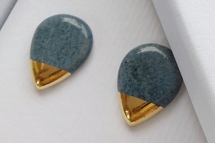 A pair of ceramic teardrop-shaped earrings with a speckled slate-blue top half and a polished gold V-shaped bottom half, displayed inside a white jewelry box.
