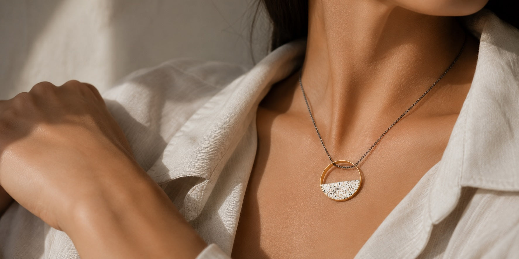 A close-up lifestyle shot of a woman wearing a delicate gold necklace with a half-circle pendant featuring a terrazzo inlay made from tiny fragments of natural stone in white and silver tones. She wears a white linen shirt in warm Mediterranean sunlight.